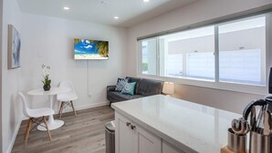 A modern kitchen with a white countertop, a dining area with white chairs, a sofa with cushions, and a TV mounted on the wall.