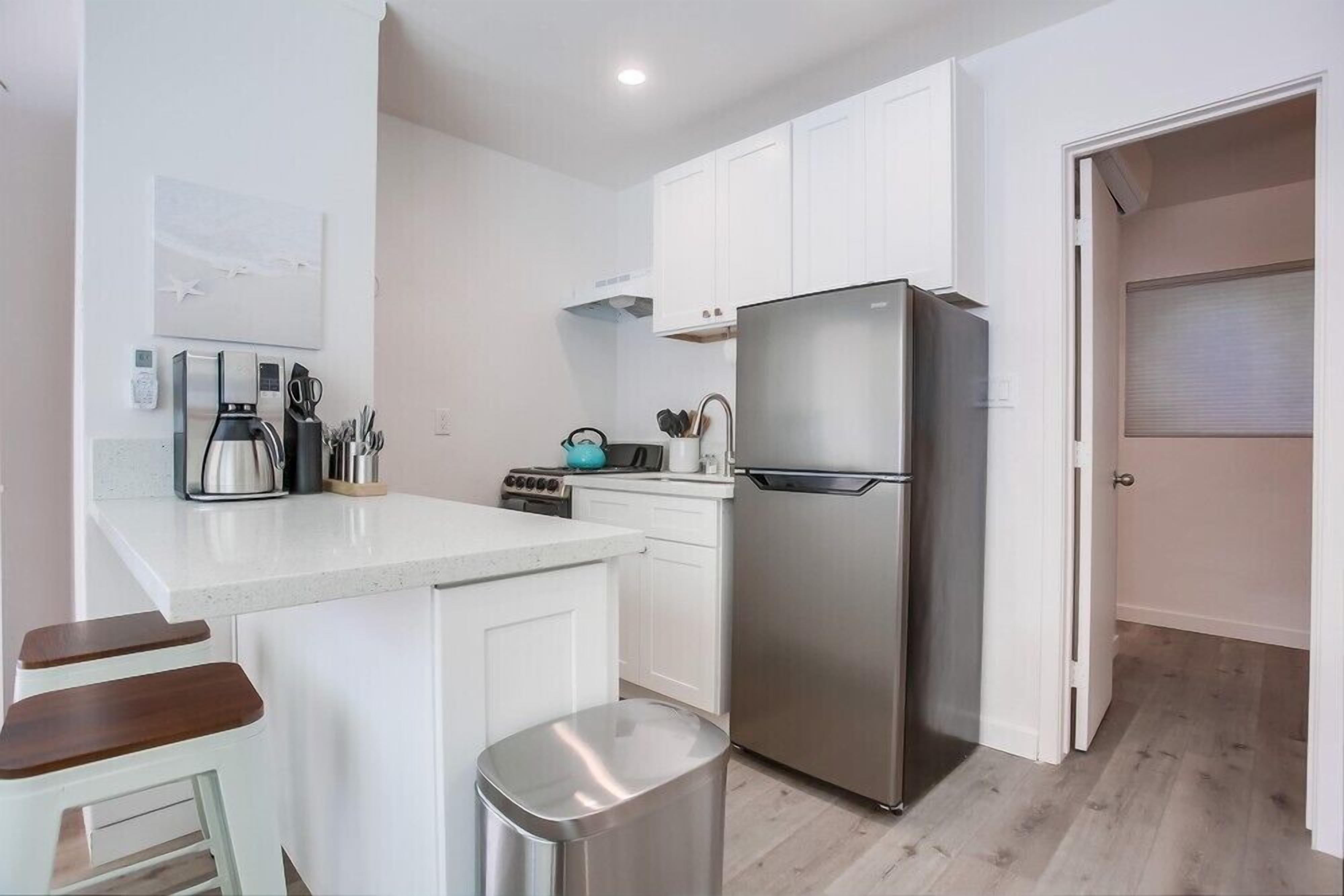 A modern kitchen with white cabinets, a stainless steel refrigerator, a coffee maker, and a toaster. There are two wooden stools and a trash bin.