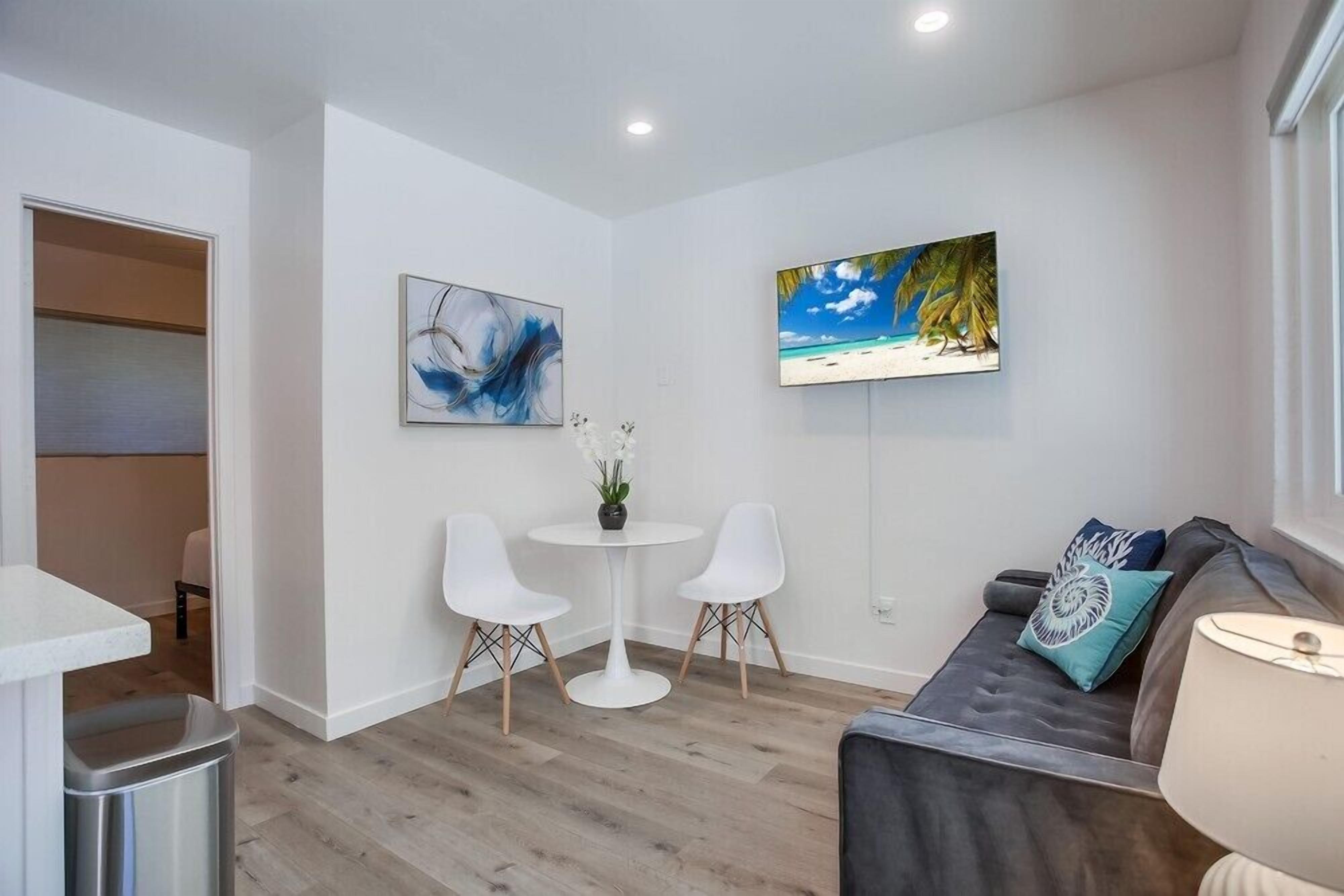 Een moderne woonkamer met een grijze bank, een ronde witte tafel en twee witte stoelen. Er hangt een schilderij van een strand aan de muur en op de tafel staat een kleine pot met een plantje.