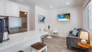 A modern kitchen with a refrigerator, a coffee maker, and a bar stool. There is a dining area with a round table and chairs. A sofa with a lamp and decorative pillows is against the wall.