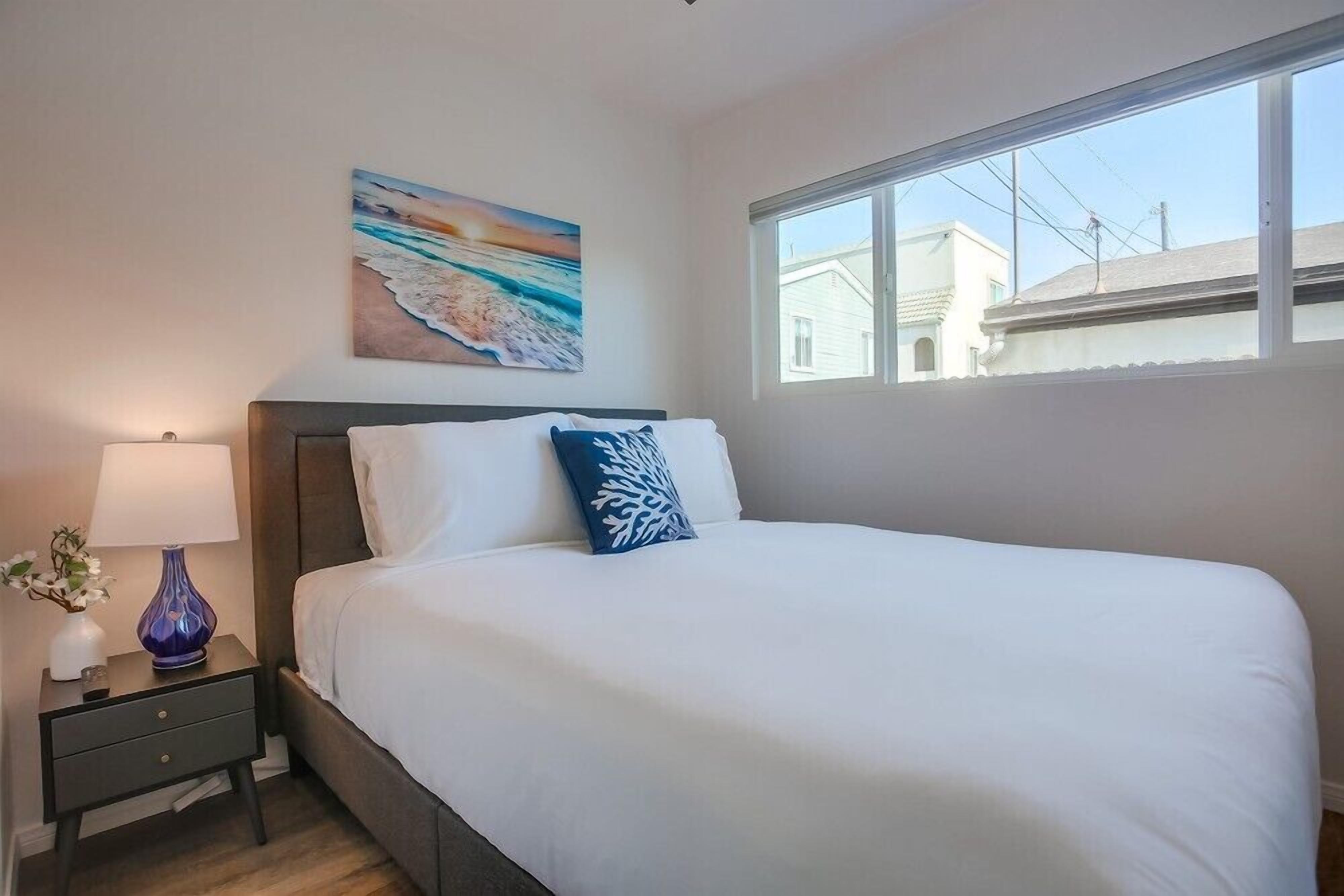 Une chambre à coucher avec un lit, une table de chevet, une lampe, un tableau et une fenêtre.