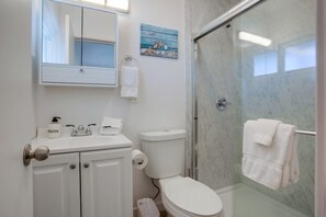 Un baño con lavamanos blanco, inodoro y ducha con cerramiento de vidrio. Se ve un perchero con una toalla y un pequeño adorno en la pared.