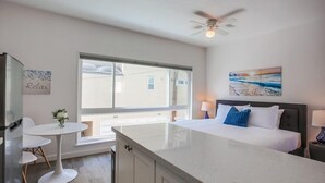 Ett modernt hotellrum med en stor säng, ett litet matbord, ett köksutrymme och ett stort fönster med utsikt över omgivningen.