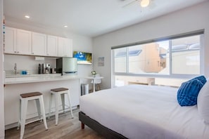 A modern hotel room with a large bed, a small kitchenette, a dining area, and a view of the city.