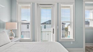 Un dormitorio con cama, una mesita de noche con lámpara y vistas a través de ventanas con marco blanco.
