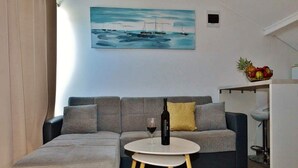 A modern living room with a grey sectional sofa, a small round table with a wine bottle and glass, and a painting of boats on the wall. - Apartments Brzica (Novi Vinodolski)