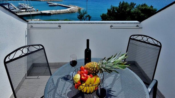 A balcony with a table set for two, featuring a fruit bowl, a bottle, and a glass of red wine. The view includes a marina and a body of water with distant mountains. - Apartments Brzica (Novi Vinodolski)
