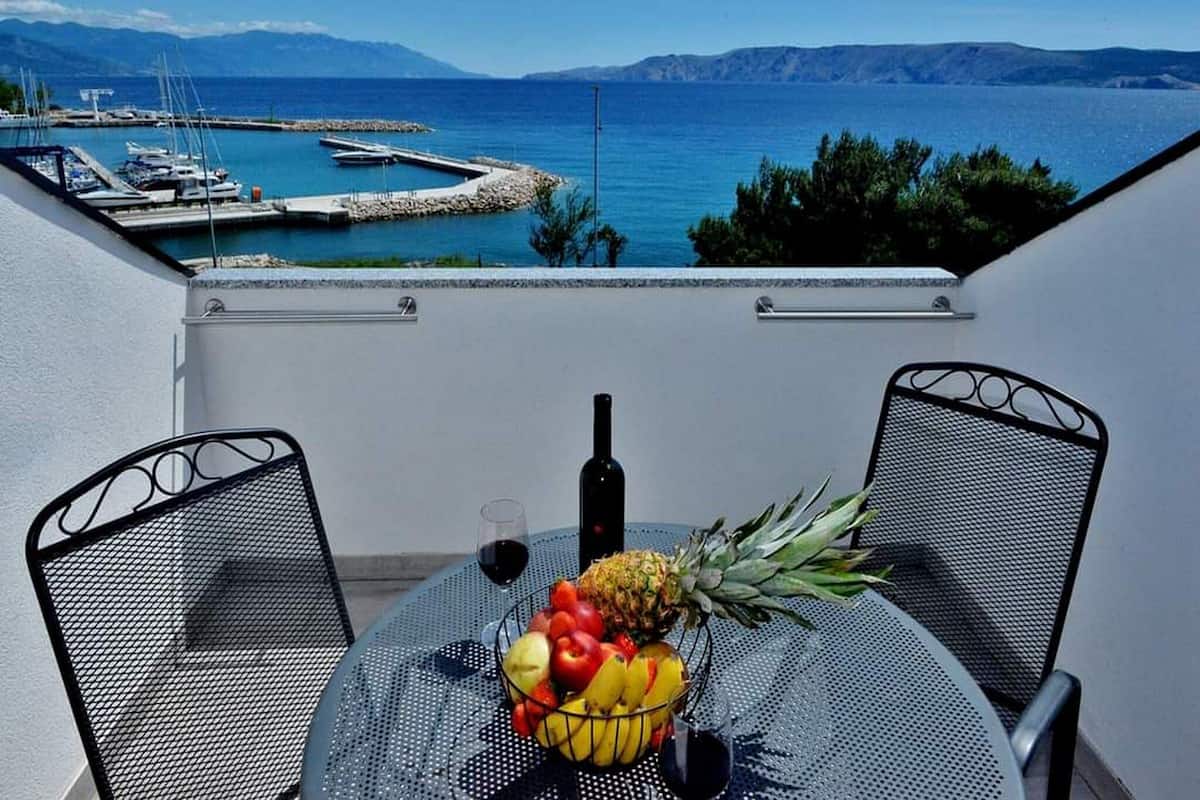 Un balcon avec une table dressée pour deux personnes, agrémentée d’un panier de fruits, d’une bouteille et d’un verre de vin rouge. La vue comprend une marina et une étendue d’eau, avec des montagnes qui se dessinent à l’horizon.