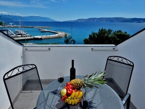 Ein Balkon mit Tisch für zwei Personen, ausgestattet mit einer Obstschale, einer Flasche und einem Glas Rotwein. Im Hintergrund ist eine Marina und ein Gewässer mit entfernten Bergen zu sehen.