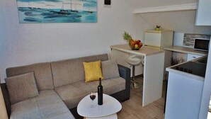 A modern living space with a sofa, a small round table with a bottle and glass, a kitchen area with a fruit bowl, and a painting of a seascape on the wall. - Apartments Brzica (Novi Vinodolski)