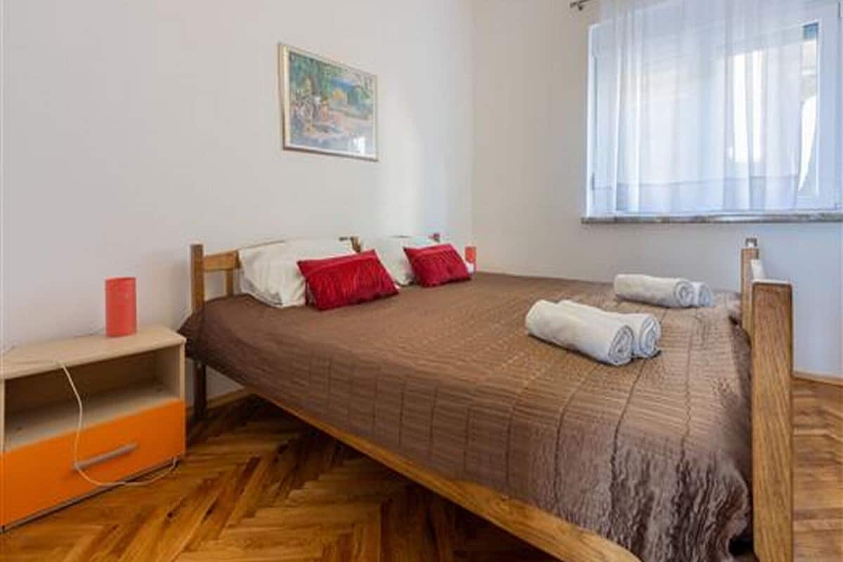 Un lit simple avec un cadre en bois, un oreiller rouge et un oreiller blanc. Il y a un tableau au mur et une fenêtre avec des rideaux légers.