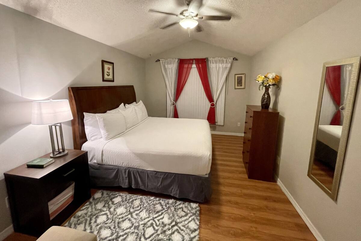 A bedroom with a bed, a bedside table with a lamp, a dresser, a ceiling fan, and a mirror.