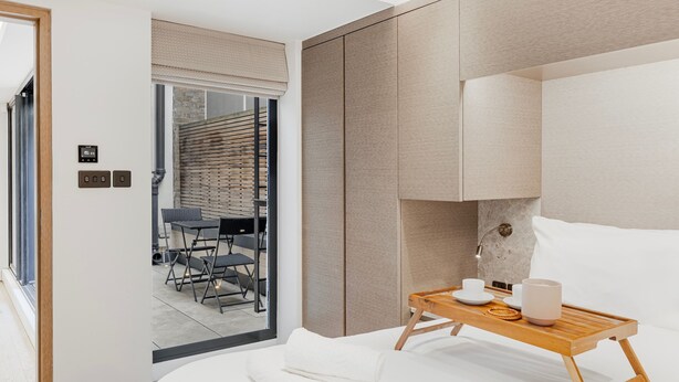 A modern hotel room with a bed, a wooden tray with cups and a bowl, a window with blinds, and a view of an outdoor seating area.