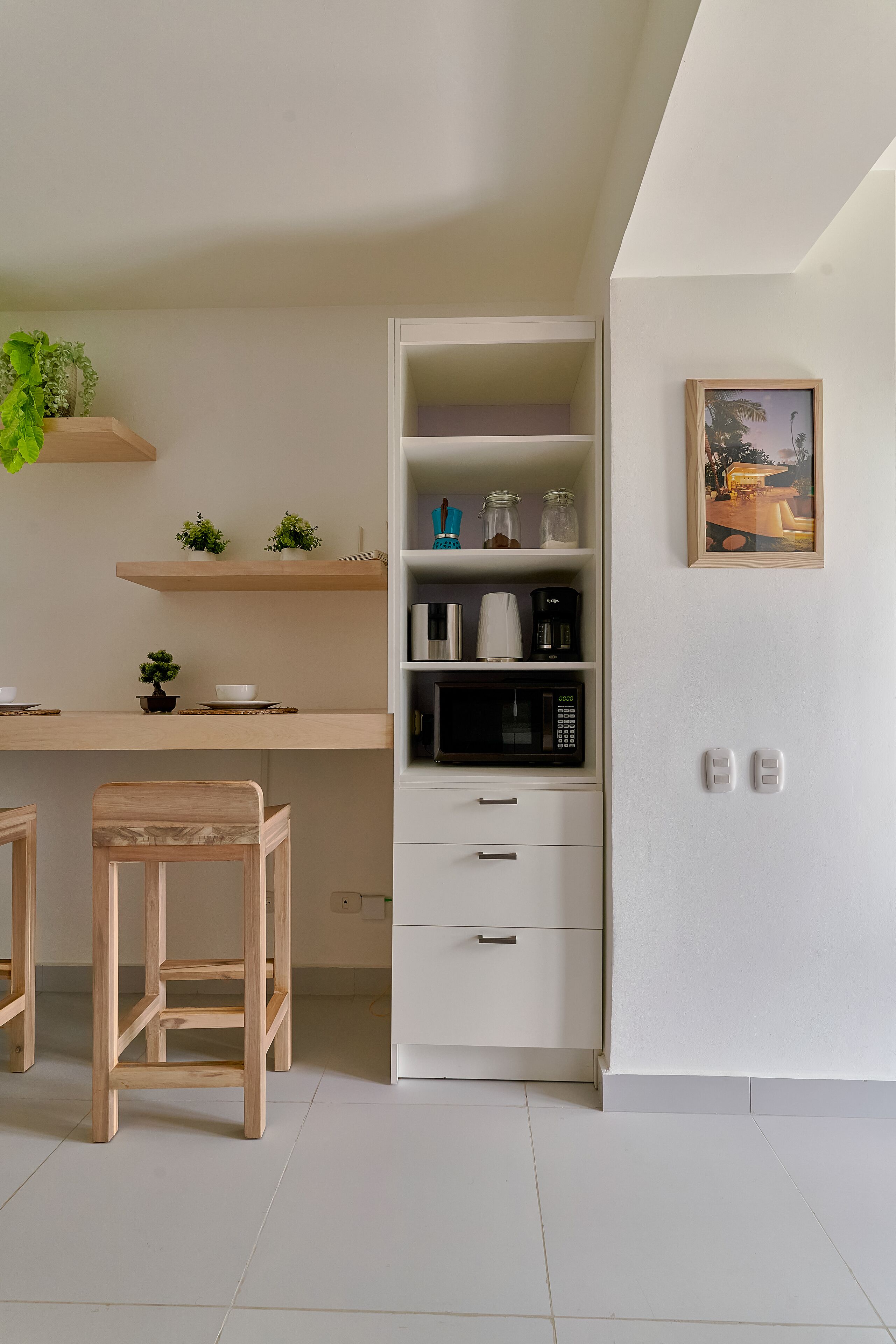 Cocina moderna con armarios blancos, microondas y cafetera. Hay dos taburetes de madera y una pequeña planta en maceta sobre la encimera.