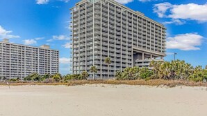 Un edificio alto con múltiples balcones, cerca de una playa arenosa y vegetación escasa.