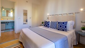 A bedroom with a large bed, a nightstand, a bedside table, a mirror, and a bathroom visible through an open door. - Borgo 66 in Alghero With 2 Bedrooms and 2 Bathrooms (Alghero)