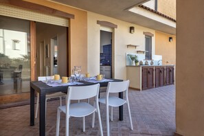 An outdoor dining area with a table set for two, white chairs, and a kitchen area with cabinets and a sink.