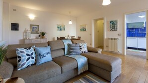 A cozy living room with a sofa, a dining table, and a kitchen area visible through an open door. - Borgo 66 in Alghero With 2 Bedrooms and 2 Bathrooms (Alghero)