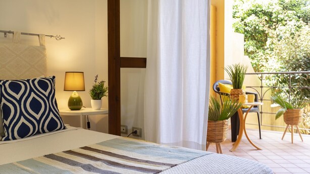 A hotel room with a bed, a bedside table with a lamp, a potted plant, and a view of the outdoor balcony with chairs and plants. - Borgo 66 in Alghero With 2 Bedrooms and 2 Bathrooms (Alghero)