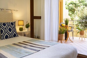 A hotel room with a bed, a bedside table with a lamp, a potted plant, and a view of the outdoor balcony with chairs and plants.