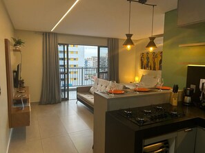 A modern kitchen with a central island, a dining area with a sofa, and a balcony with a city view. - Bela Hospedagem - Lindo flat no Box 150 (João Pessoa)