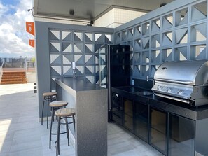 An outdoor kitchen with a grill, bar stools, and a refrigerator. - Bela Hospedagem - Lindo flat no Box 150 (João Pessoa)