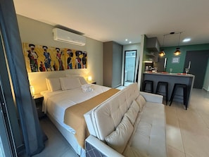 Quarto de hotel com cama, sofá, área de refeições com banquetas, ar condicionado e um quadro na parede.