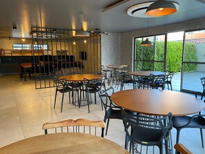 A modern cafe with round tables and black chairs, large windows with a view of greenery, and pendant lighting. - Bela Hospedagem - Lindo flat no Box 150 (João Pessoa)