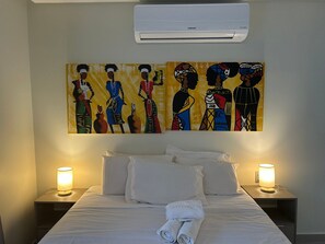 A hotel room with a bed, white pillows, a rolled towel, two bedside lamps, and a wall-mounted air conditioner. - Bela Hospedagem - Lindo flat no Box 150 (João Pessoa)