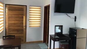 A room with a wooden door, a window with blinds, a flat-screen TV mounted on the wall, a desk with a chair, and a small table. - Tequila Cove (Panglao)