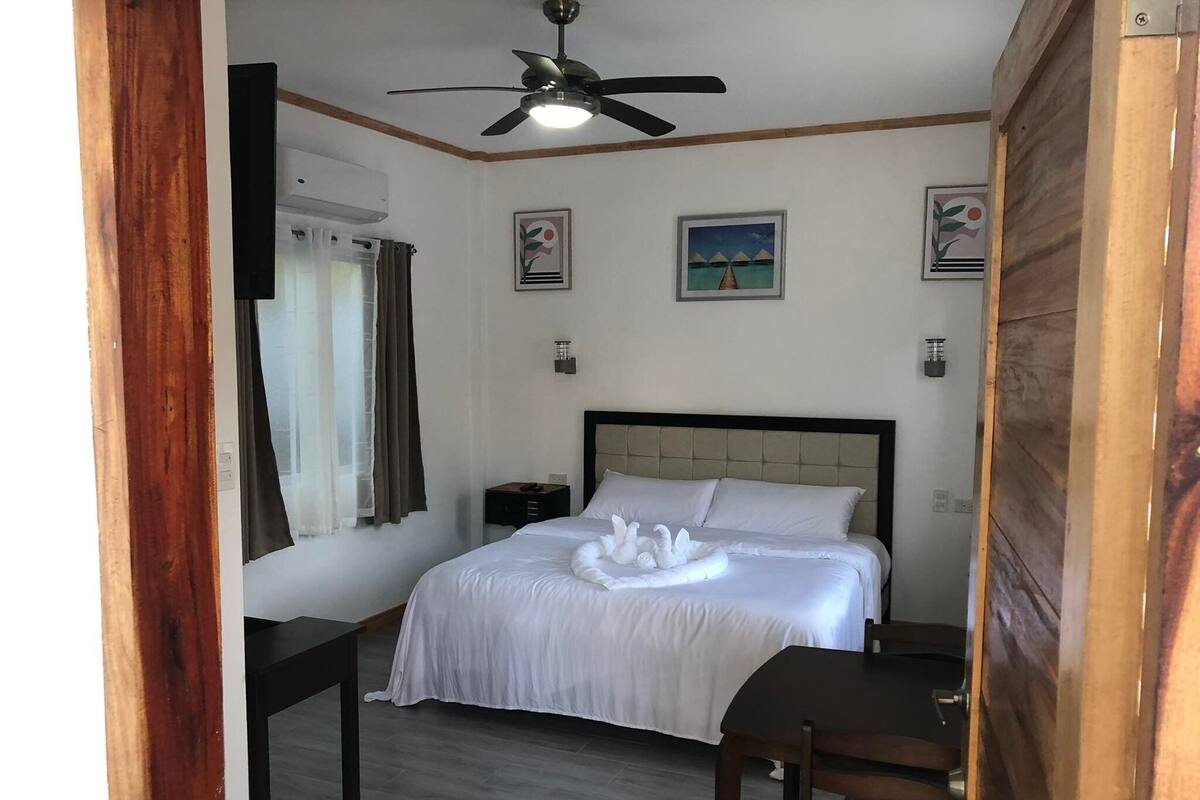 A hotel room with a bed, a desk, a chair, a ceiling fan, and artwork on the walls.