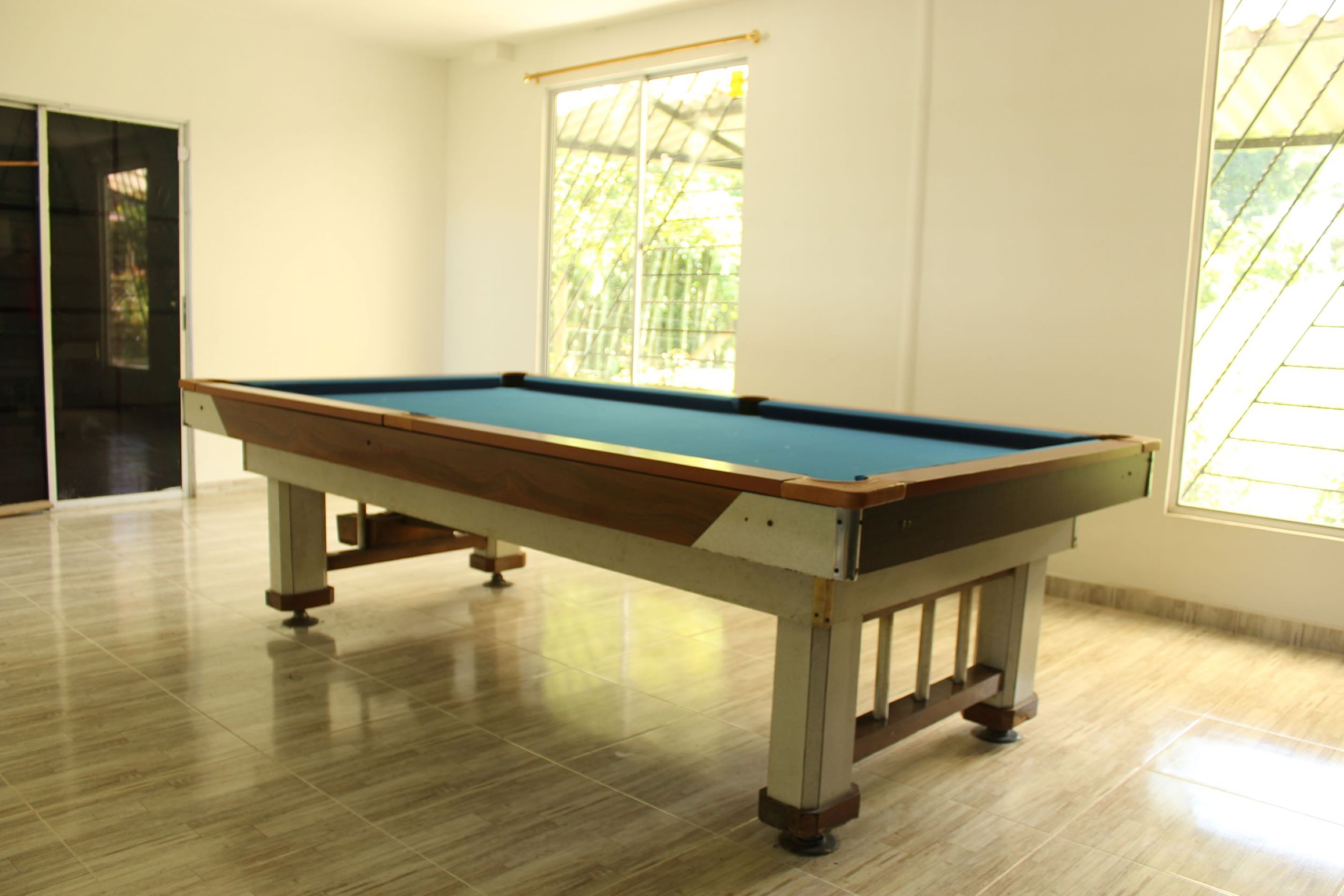 Una mesa de billar en una sala con ventanas amplias y piso de baldosas.