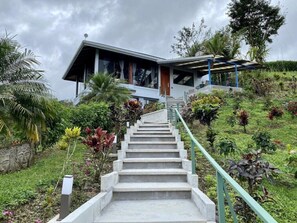 A staircase leading to a modern house with a covered porch, surrounded by lush greenery and a cloudy sky.