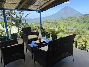 A covered outdoor seating area with wicker furniture and potted plants, overlooking a lush landscape and a mountain.