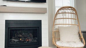 A modern living room with a fireplace, a wicker chair, and a television mounted on the wall. - Cozy Cottage in Seabrook w/ Private Hot Tub (Pacific Beach)