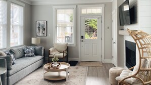 A living room with a grey sofa, a round coffee table, a wicker chair, and a television mounted on the wall. - Cozy Cottage in Seabrook w/ Private Hot Tub (Pacific Beach)