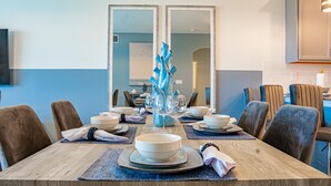 Un comedor con una mesa de madera dispuesta para seis personas, rodeada de sillas. Hay dos espejos en la pared, un jarrón con detalles decorativos azules, y un televisor de pantalla plana montado en la pared.
