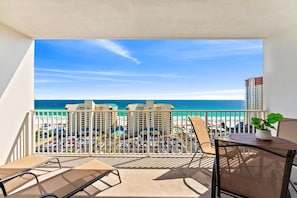 Ein Balkon mit Blick auf Strand und Meer, ein Stuhl, ein Tisch mit einer Pflanze und ein Parkplatz darunter.