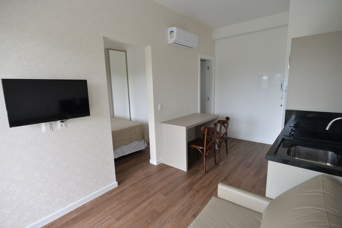 A compact living space with a kitchenette, a flat-screen TV mounted on the wall, a dining area with a table and chairs, and a bed in the adjacent room.