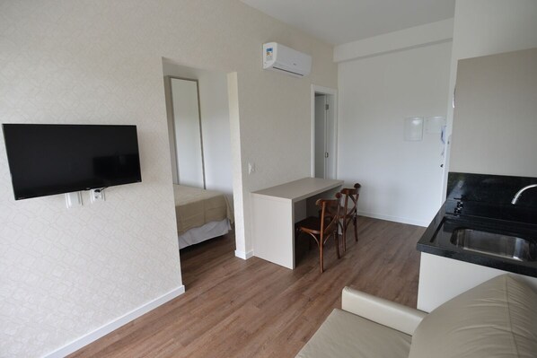 A compact living space with a kitchenette, a flat-screen TV mounted on the wall, a dining area with a table and chairs, and a bed in the adjacent room. - Vila do Centro Apart Hotel (Bombinhas)