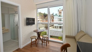 A modern living room with a sofa, a TV mounted on the wall, a glass door leading to a balcony, and a small round table with two stools. - Vila do Centro Apart Hotel (Bombinhas)