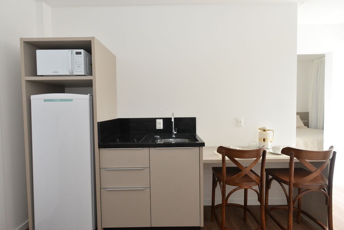 A compact kitchen with a microwave, refrigerator, and a small sink. There are two wooden chairs and a white table with a coffee maker.