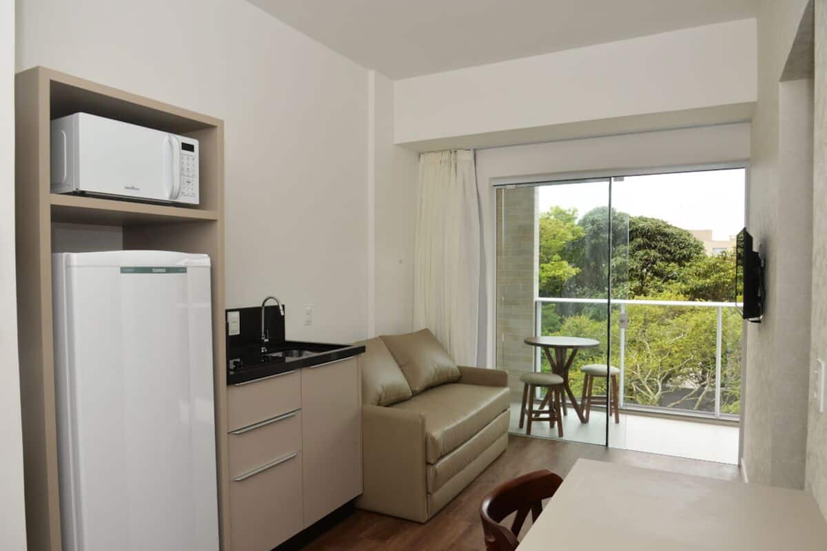 Een functionele keuken met een magnetron, koelkast en wastafel. Een beigekleurige bank en een eettafel met stoelen bevinden zich in de woonruimte. Een balkon met een glazen deur en uitzicht op groen is zichtbaar.