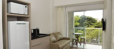 Cocina compacta con microondas, refrigerador y lavabo. Un sofá color crema y una pequeña mesa de comedor con sillas se encuentran en la sala. Se puede apreciar un balcón con puerta de vidrio y vistas a zonas verdes.