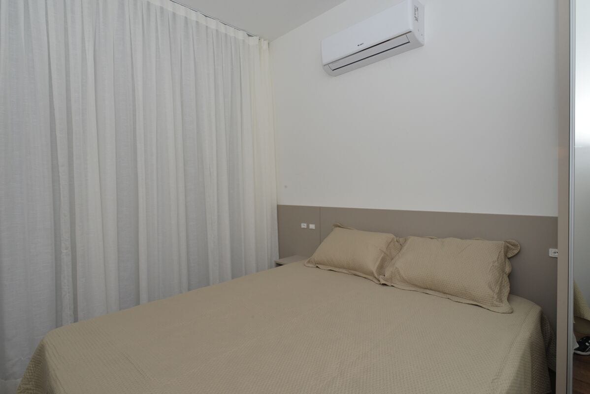 A hotel room with a bed, beige bedding, a white curtain, and an air conditioning unit on the wall.