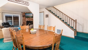 A dining area with a wooden table and chairs, a brick fireplace, and a staircase with teal carpet. - Snow Summit Getaway - 2-bedroom in Big Bear Lake, Walk to Slopes, Cable TV, Deck, BBQ, Views (Big Bear Lake)