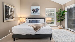 Une chambre à coucher avec un grand lit, une fenêtre avec des stores, une lampe fixée au mur et un tableau encadré représentant un bison.