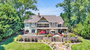 A two-story house with a large deck, outdoor seating, and a stone retaining wall. - Beautiful lakeside family or corporate retreat with pool, hot tub, and  more! (Huddleston)