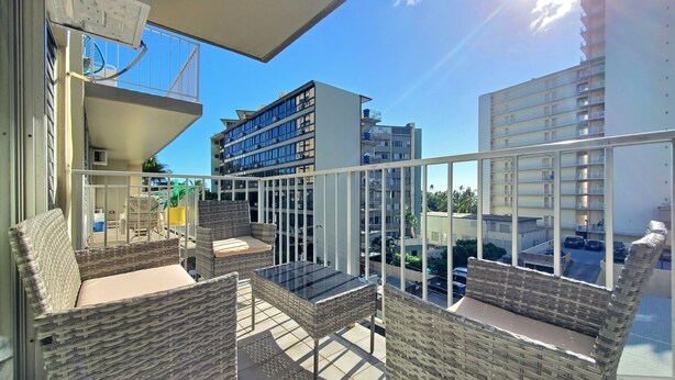 Een balkon met rieten meubels en een glazen tafel, met uitzicht op een parkeerterrein en gebouwen.
