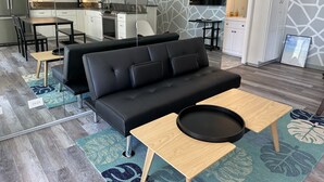 Een moderne woonkamer met een zwarte bank, een houten salontafel en een keuken op de achtergrond.